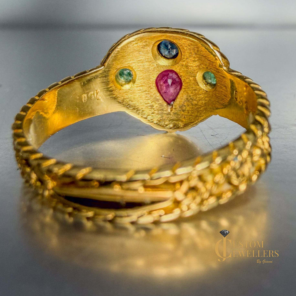 A gold ring with a textured oval top featuring a central pink gemstone and three smaller green and blue gemstones, displayed on a reflective gray surface.