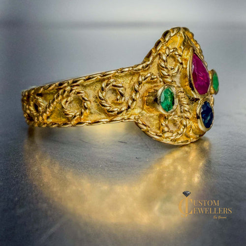 An ornate gold ring with carved floral details and colorful gemstones in pink, green, and blue, displayed on a reflective metallic surface.