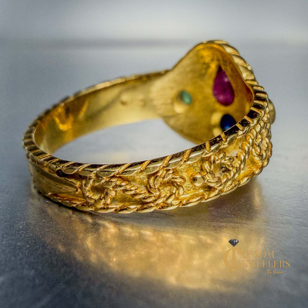 A gold bangle bracelet with intricate floral engravings and a heart-shaped end decorated with small colored gemstones, shown on a reflective gray surface.