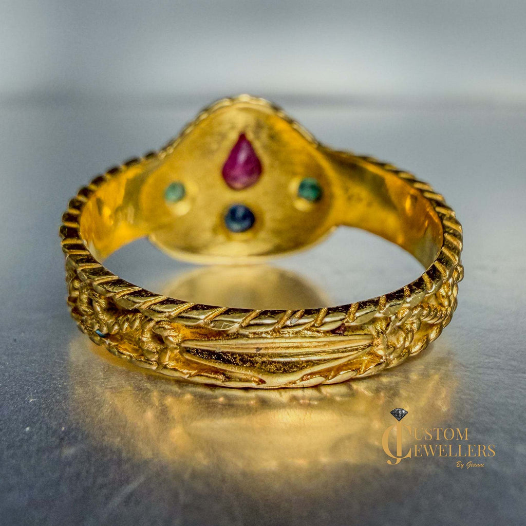 An ornate gold ring with a textured, rope-like band and a decorative top featuring a central purple teardrop gemstone and smaller blue and green stones, displayed on a reflective metallic surface.