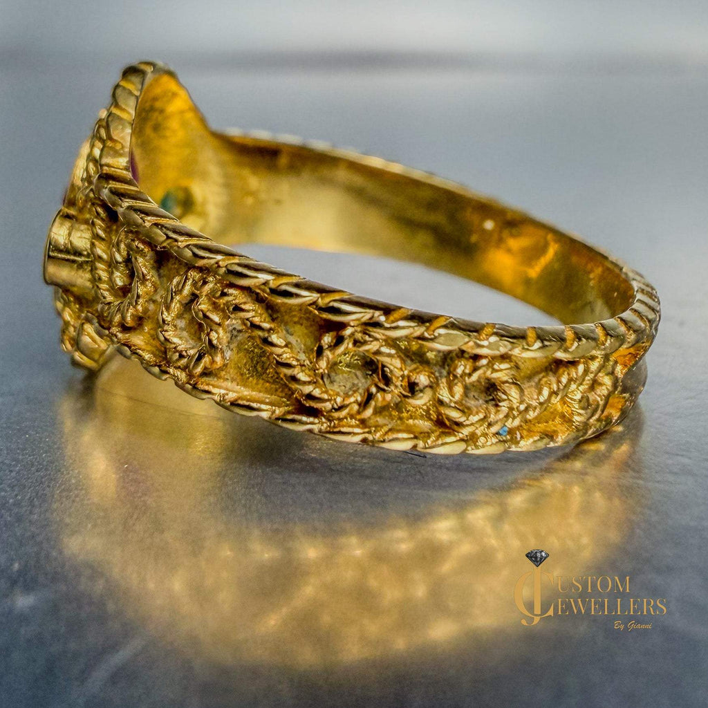 An ornate gold ring with intricate floral and leafy engravings, displayed on a reflective metallic surface.