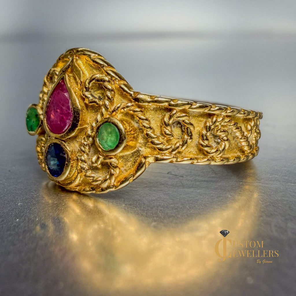 An ornate gold ring with three colorful gemstones (pink, green, and blue) set in a decorative floral pattern, displayed on a reflective gray surface.