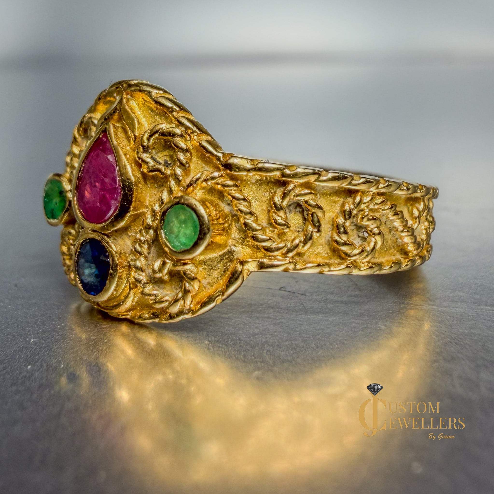 An ornate gold ring with three colorful gemstones (pink, green, and blue) set in a decorative floral pattern, displayed on a reflective gray surface.