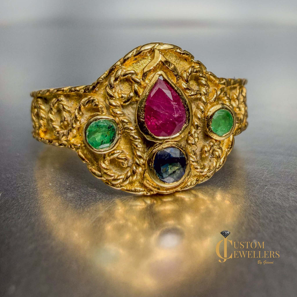 An ornate gold ring with a large central teardrop ruby gemstone and two smaller green and blue gemstones, displayed on a reflective metallic surface.