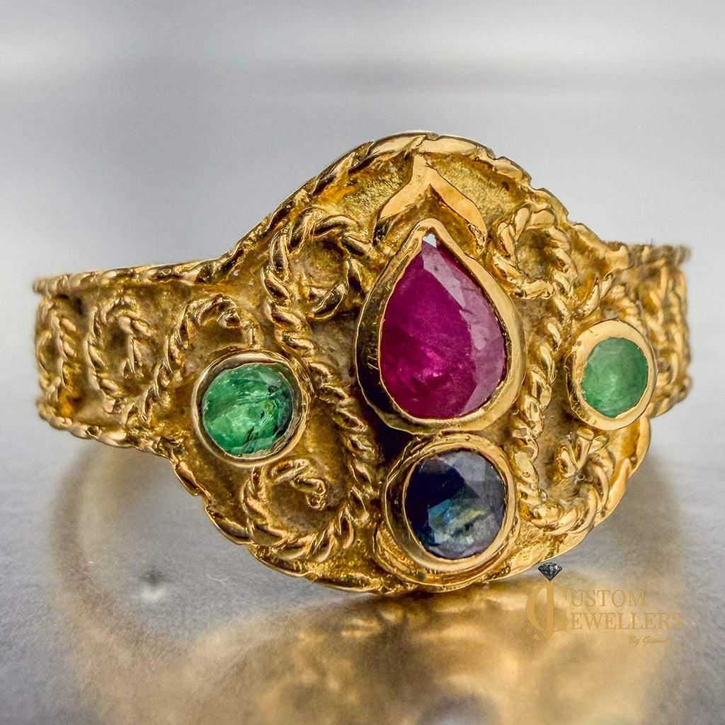 An ornate gold ring with three colored gemstones, featuring a large teardrop-shaped pinkish-red center stone, a green emerald on the left, and a dark blue oval stone on the right, set against a reflective metallic background.