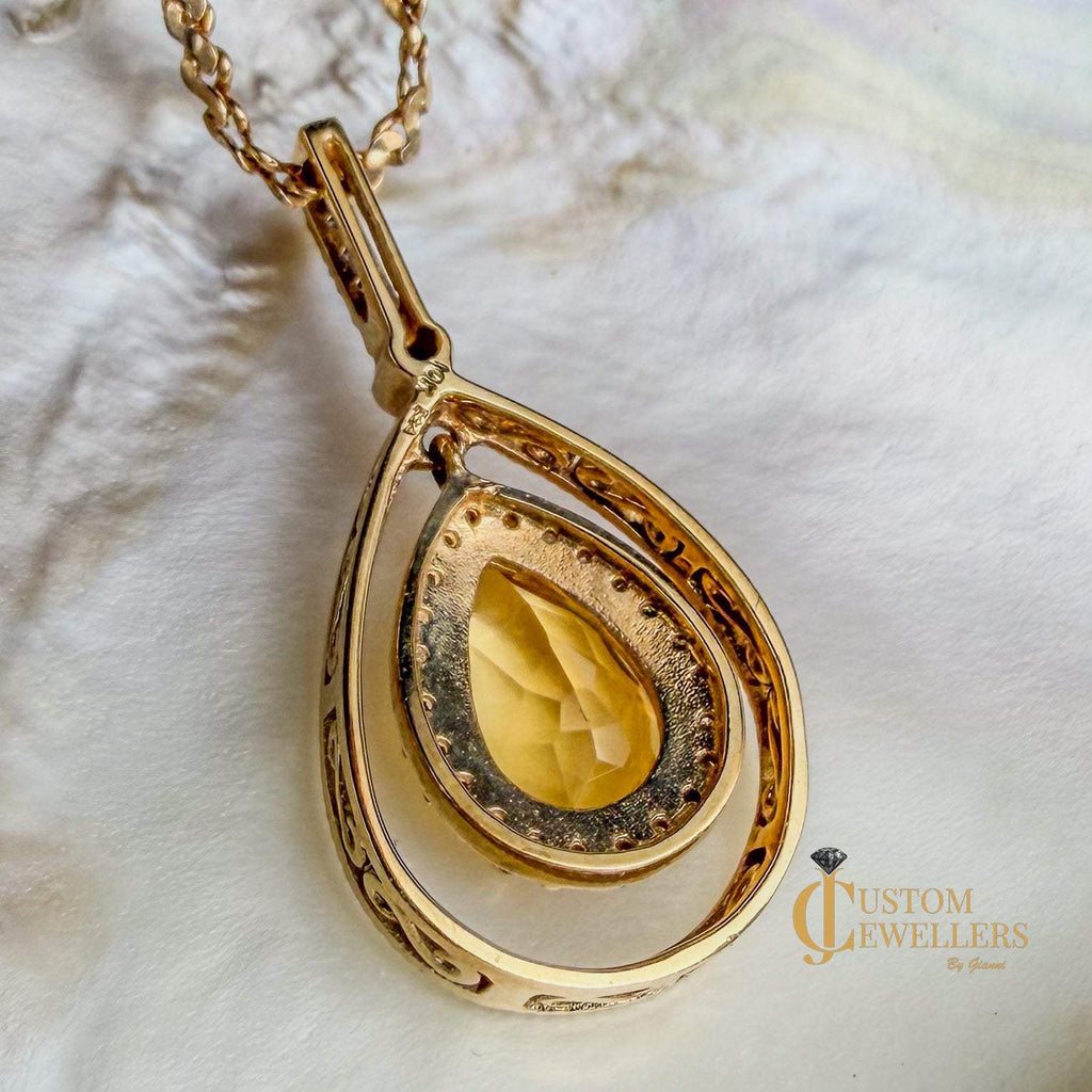 A gold teardrop pendant necklace featuring a central faceted yellow gemstone and ornate filigree detailing, displayed on a textured shell background.