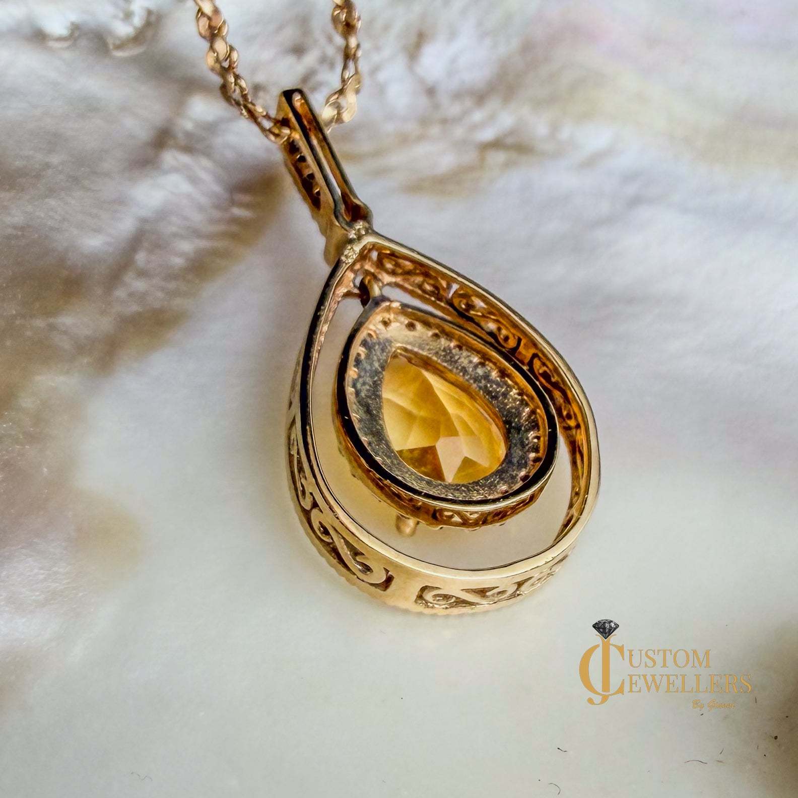 A gold teardrop pendant necklace with an ornate filigree design and a faceted yellow gemstone, resting on a white textured surface.