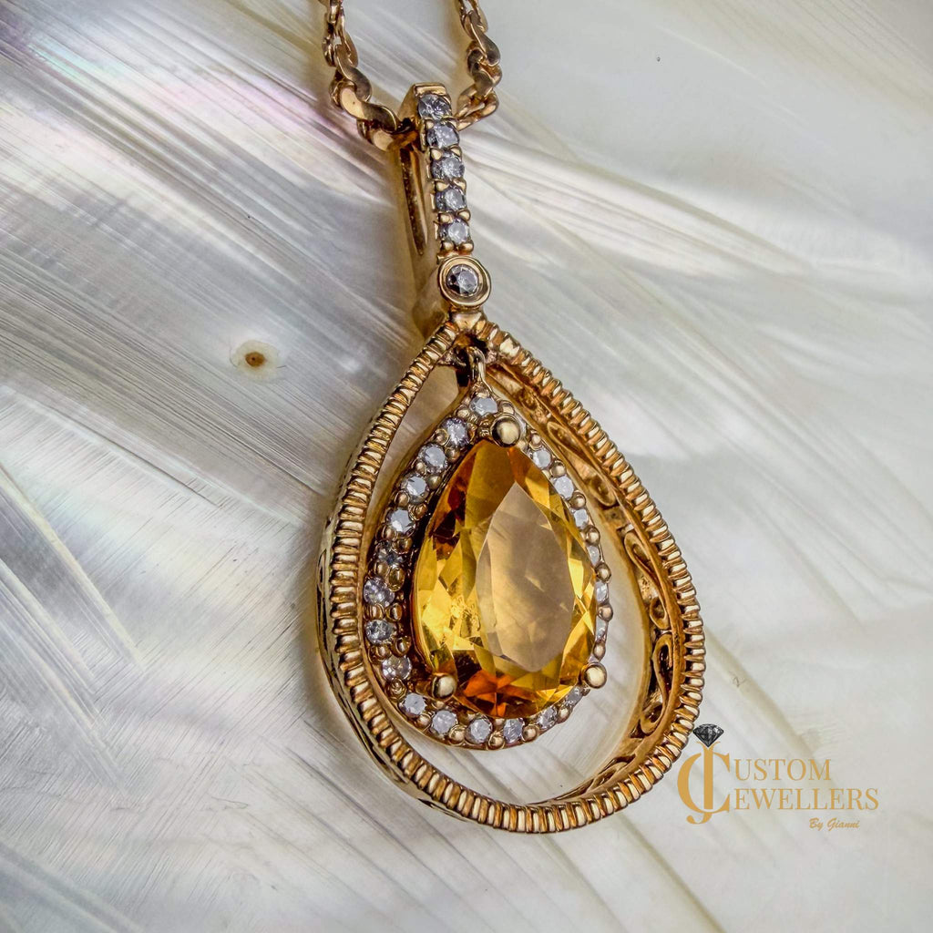 A gold teardrop pendant necklace featuring a large faceted yellow topaz center stone surrounded by small diamonds, set within a textured double-groove gold frame, resting on a soft white textured surface.