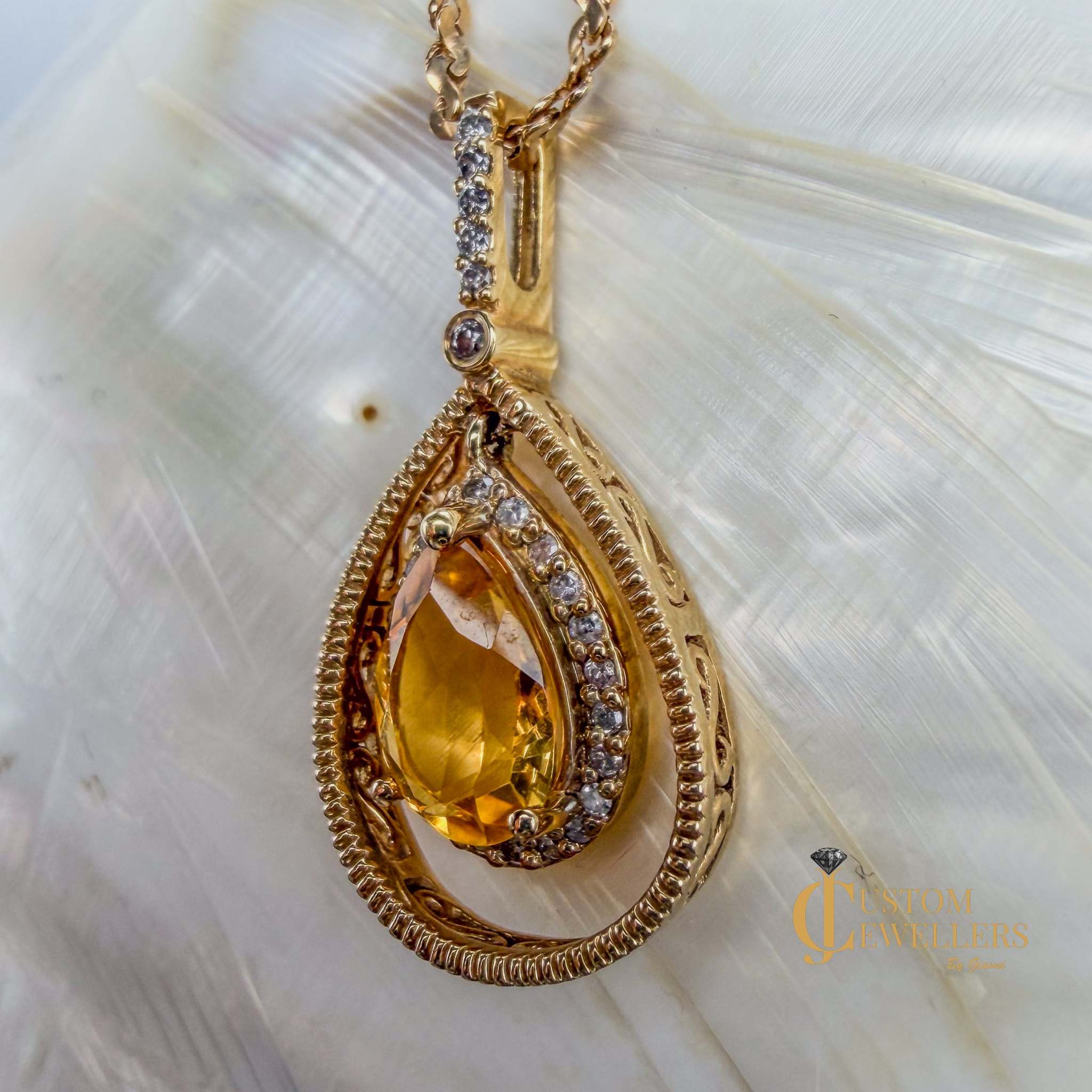 A gold teardrop pendant necklace featuring a large faceted yellow topaz center stone surrounded by small diamonds, set within a textured double-groove gold frame, resting on a soft white textured surface.