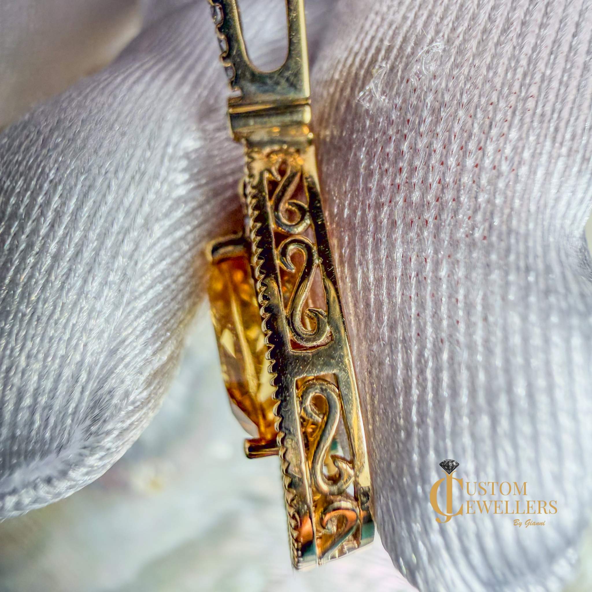 A close-up of an ornate gold-toned pendant or charm with engraved scrollwork, resting on a white textured fabric background. 