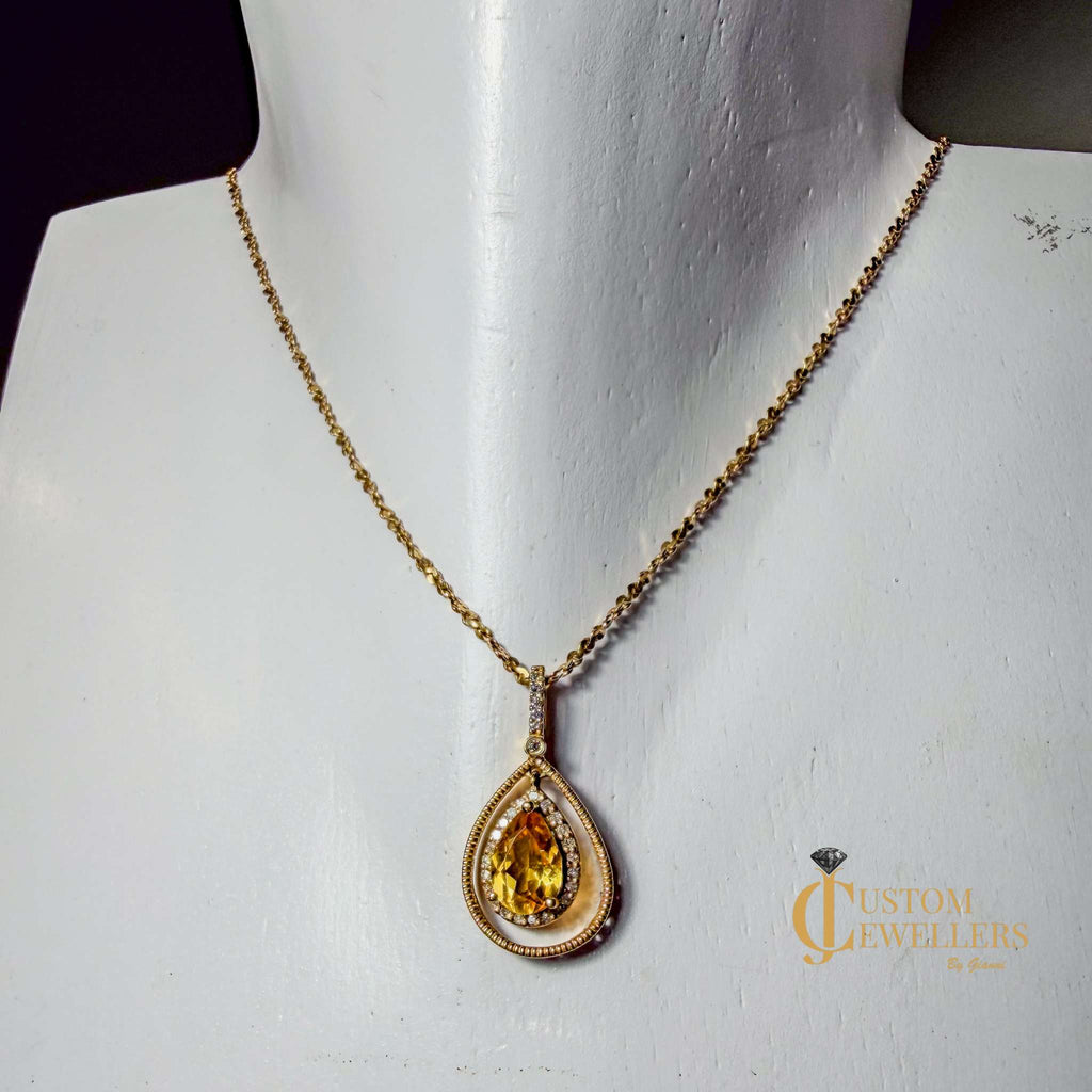 A gold teardrop pendant necklace with a yellow gemstone, displayed on a white jewelry mannequin bust. The pendant features a central yellow stone surrounded by a halo of small clear stones and a textured gold border, suspended from a delicate gold chain.