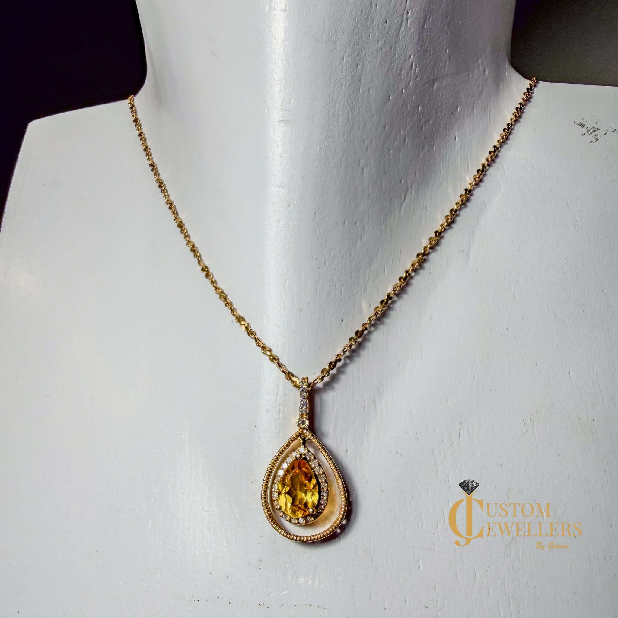 A gold teardrop pendant necklace with a yellow gemstone, displayed on a white jewelry mannequin bust. The pendant features a central yellow stone surrounded by a halo of small clear stones and a textured gold border, suspended from a delicate gold chain.