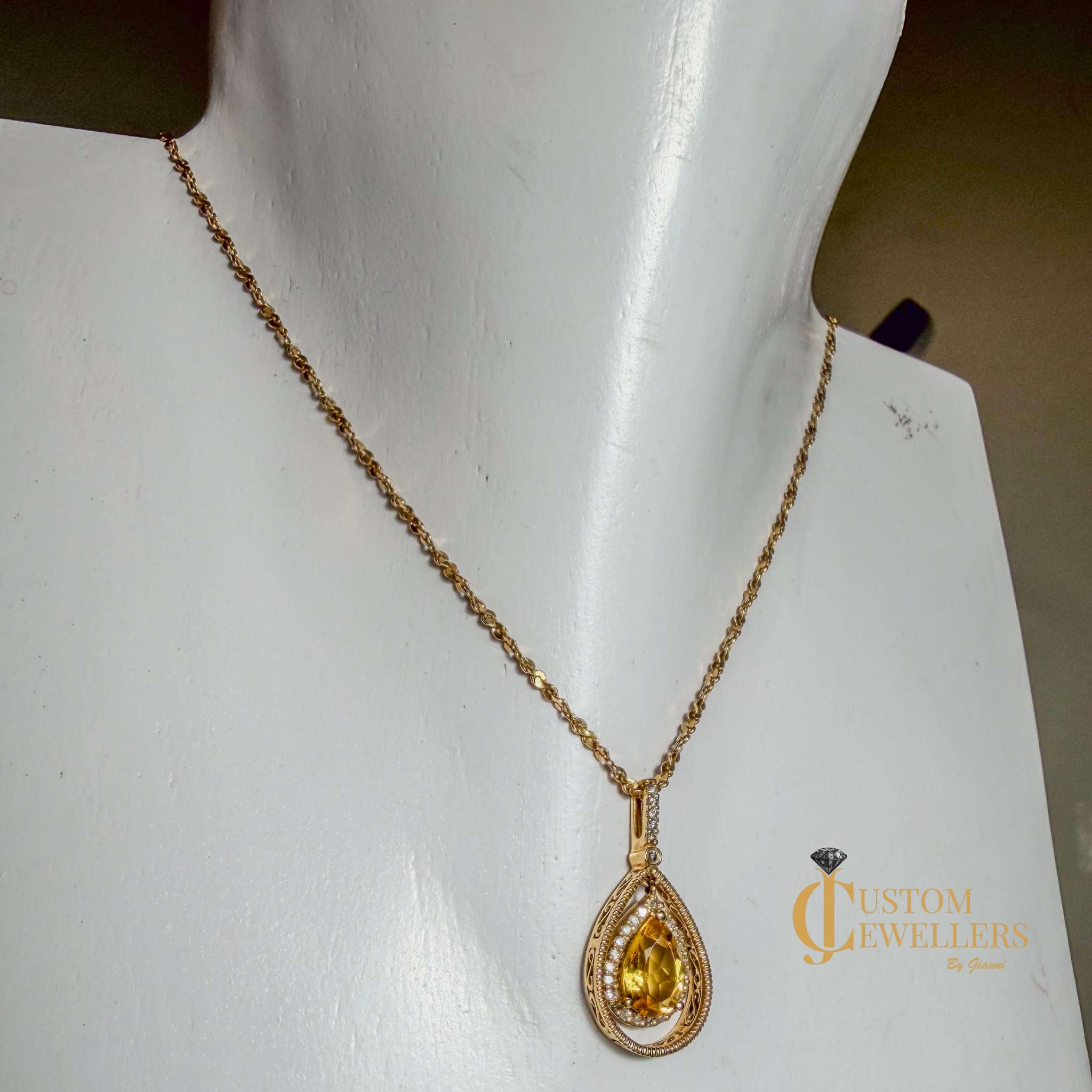 A gold teardrop pendant necklace with a yellow gemstone, displayed on a white jewelry mannequin bust. The pendant features a central yellow stone surrounded by a halo of small clear stones and a textured gold border, suspended from a delicate gold chain.