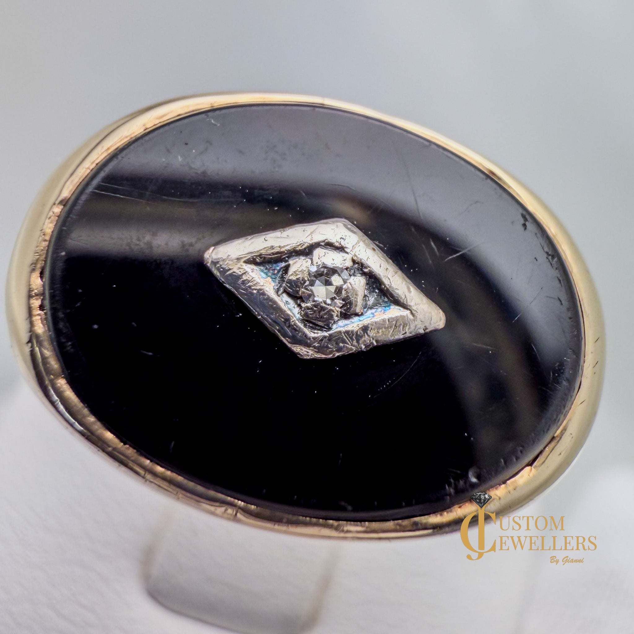 Ring with black stone and diamond center on gray background, branded 'Custom Jewellers'.