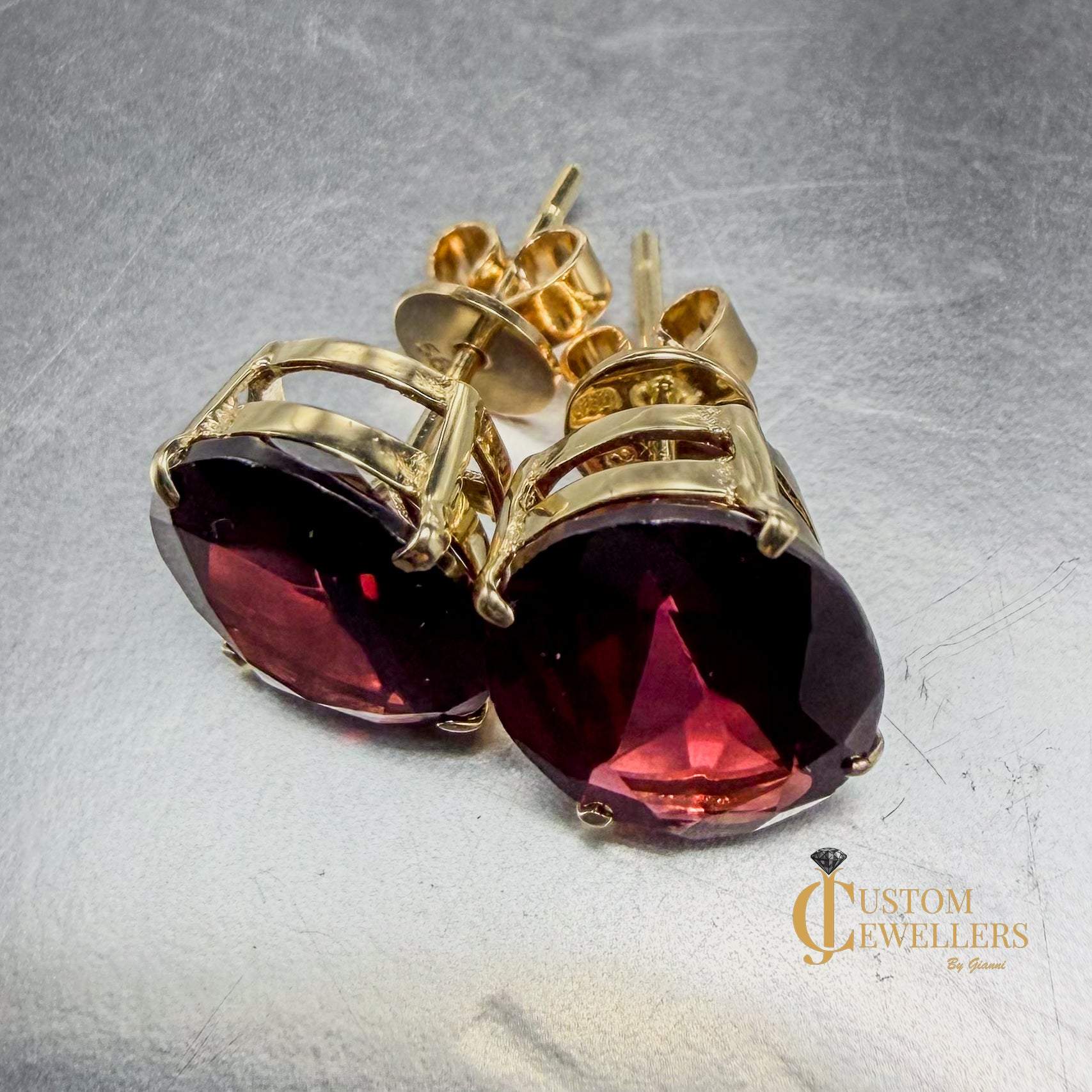 Oval faceted deep red ruby gemstone stud earrings in gold settings, shown on a reflective metallic surface with one earring facing forward and the other angled to show the side profile.