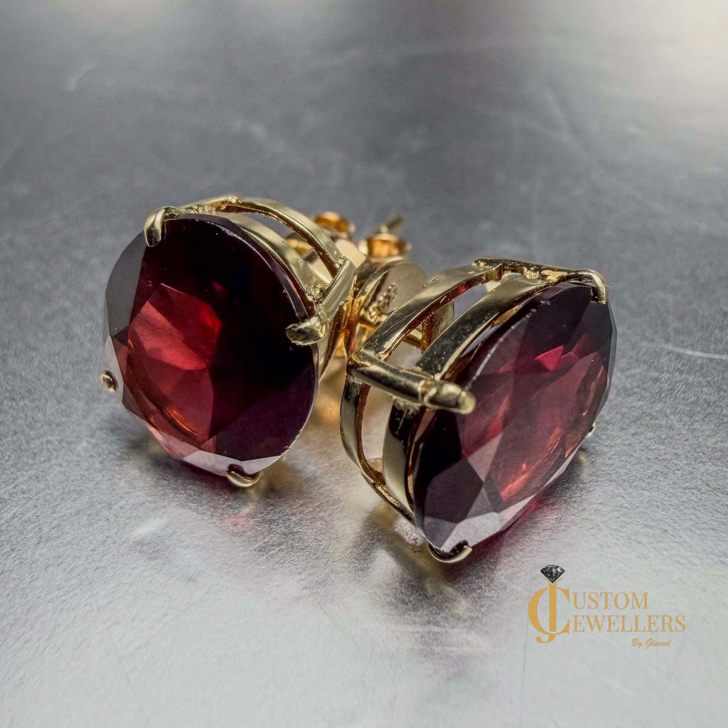 Oval faceted deep red ruby gemstone stud earrings in gold settings, shown on a reflective metallic surface with one earring facing forward and the other angled to show the side profile.