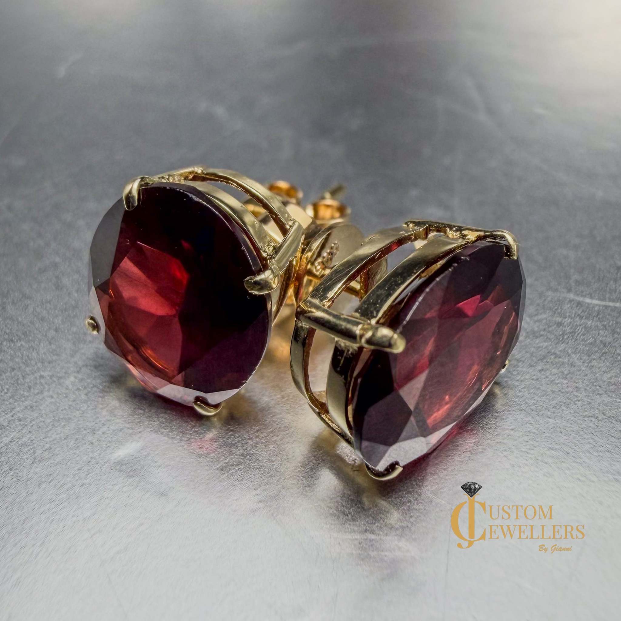 Oval faceted deep red ruby gemstone stud earrings in gold settings, shown on a reflective metallic surface with one earring facing forward and the other angled to show the side profile.