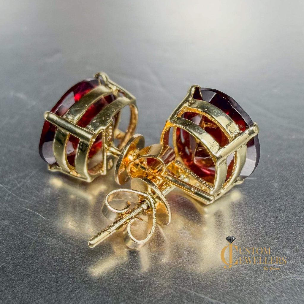 A pair of gold stud earrings with faceted red ruby gemstones, displayed on a metallic gray surface.Both earrings are turned backwards to show the post and closures.