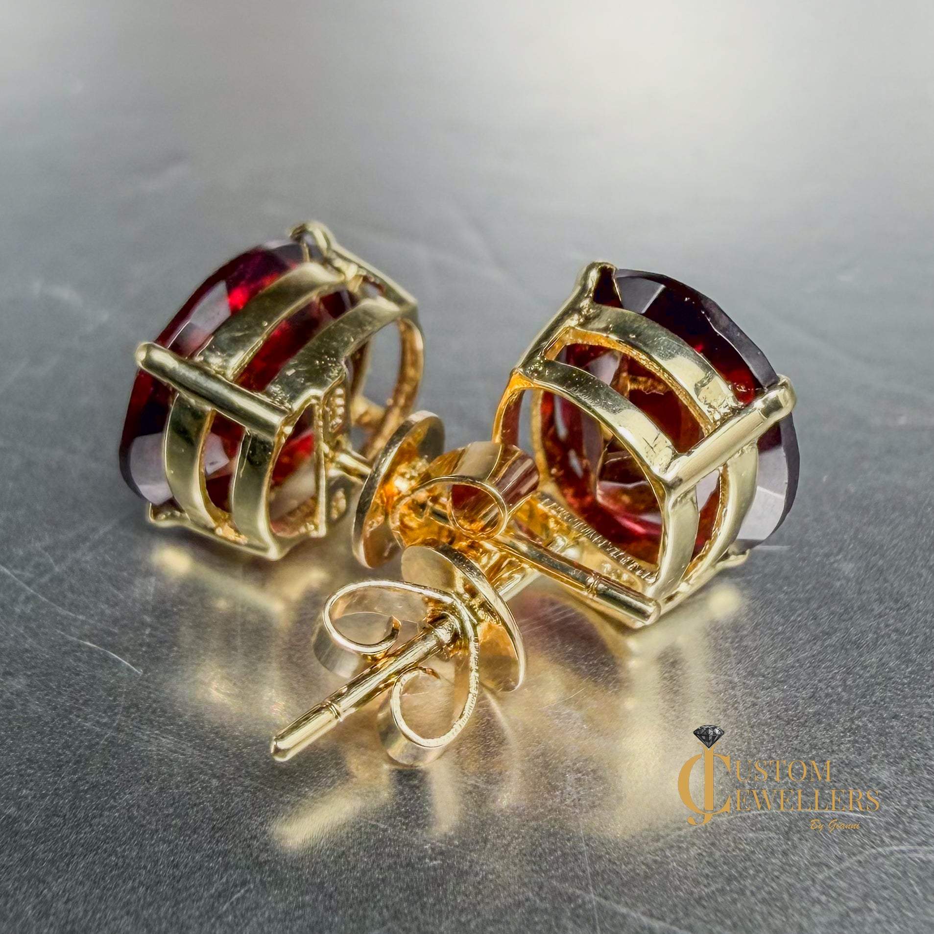 A pair of gold stud earrings with faceted red ruby gemstones, displayed on a metallic gray surface.Both earrings are turned backwards to show the post and closures.