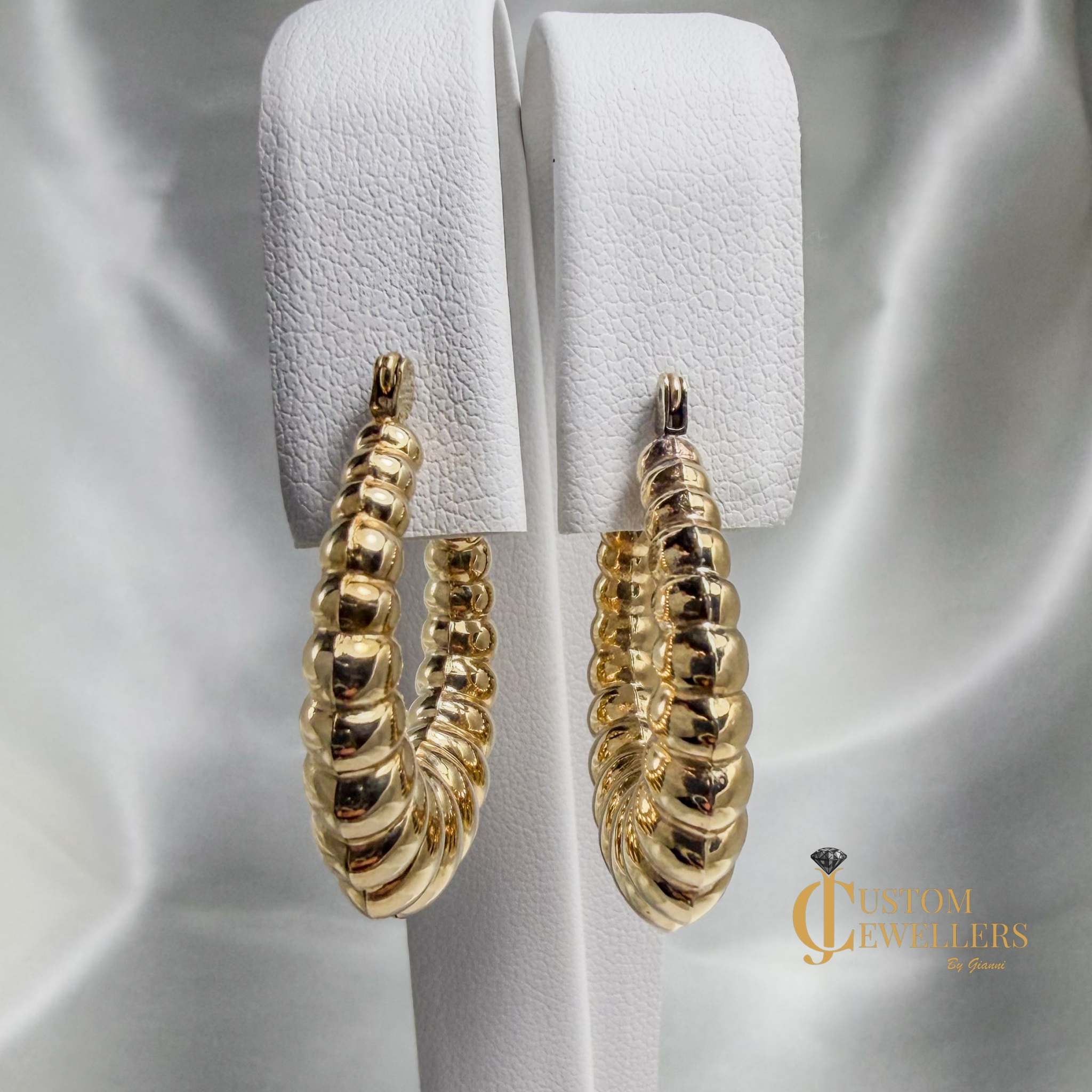 A pair of gold hoop earrings with a textured, ribbed spiral design, displayed on a white jewelry stand against a draped white satin background.