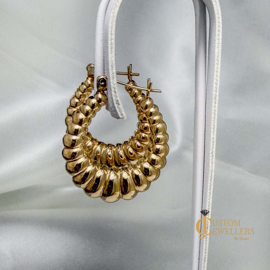 A pair of gold hoop earrings with a textured, beaded crescent design, displayed on a white fabric stand against a draped white satin background.