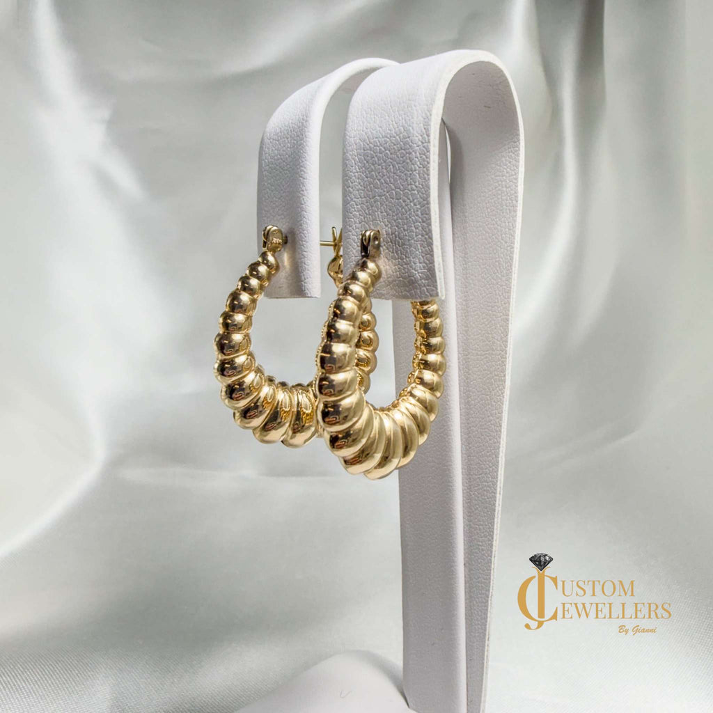 A pair of textured gold hoop earrings displayed on a white faux-leather jewelry stand against a draped white satin fabric background