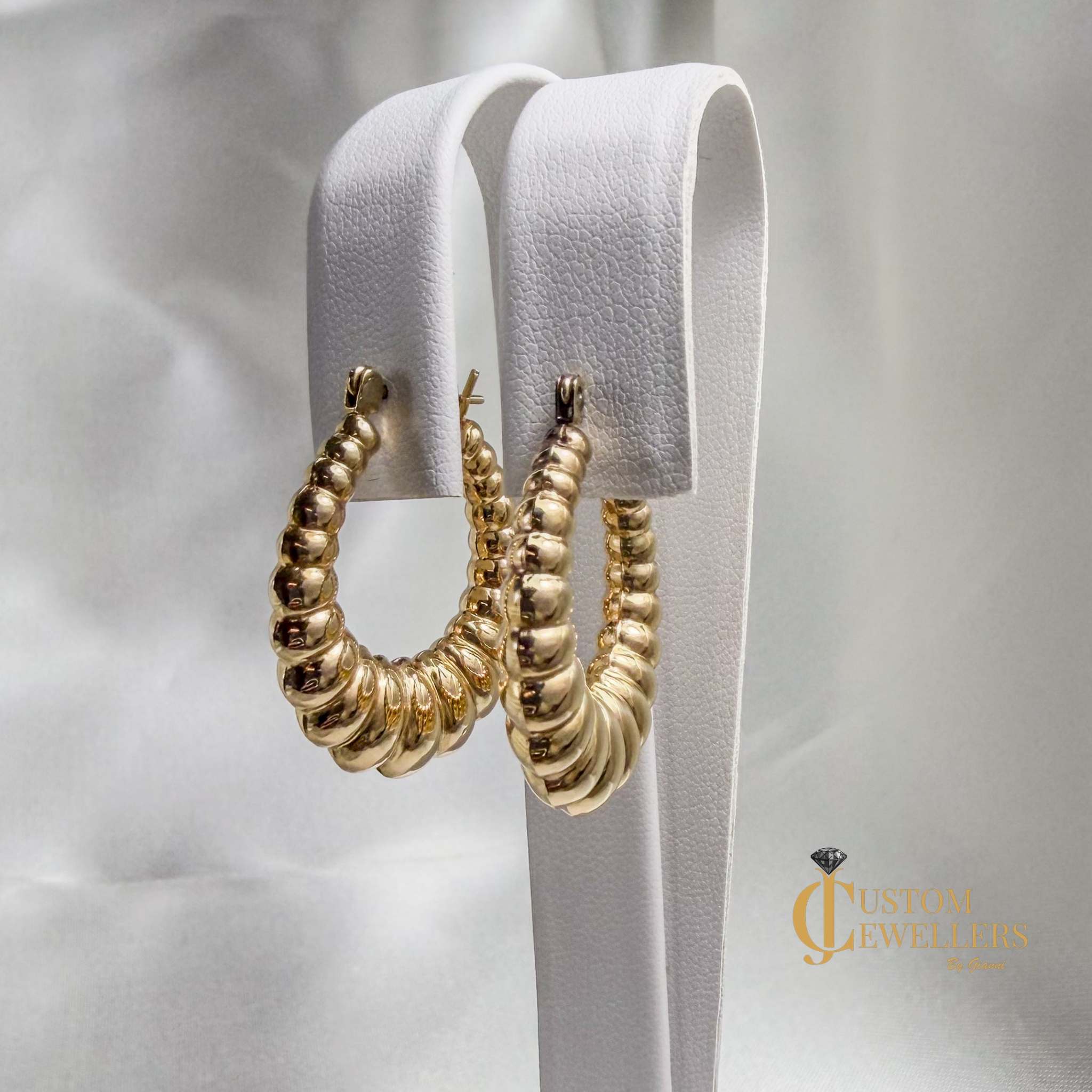 A pair of gold hoop earrings with a textured, ridged spiral design, displayed on a white textured stand against a soft white fabric background.