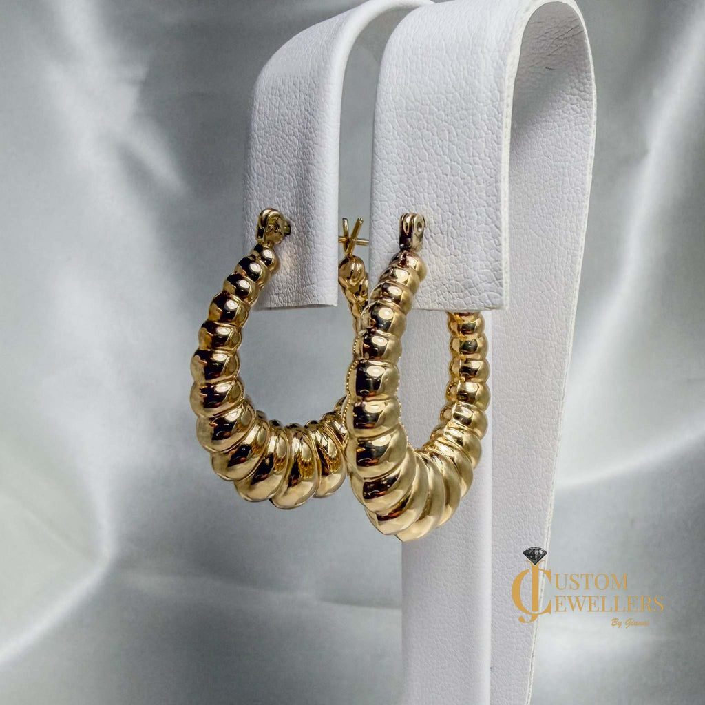 A pair of chunky, textured gold hoop earrings displayed on a white faux-leather jewelry stand against a draped silver satin background.

