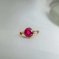 Cherie Amour Scrollwork ring in gold-tone metal with a vivid round ruby-colored center stone and ornamental side details.