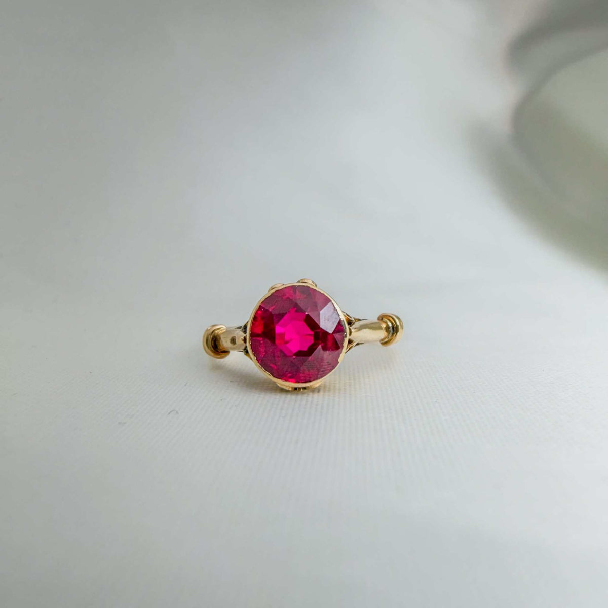Cherie Amour Scrollwork ring in gold-tone metal with a vivid round ruby-colored center stone and ornamental side details.