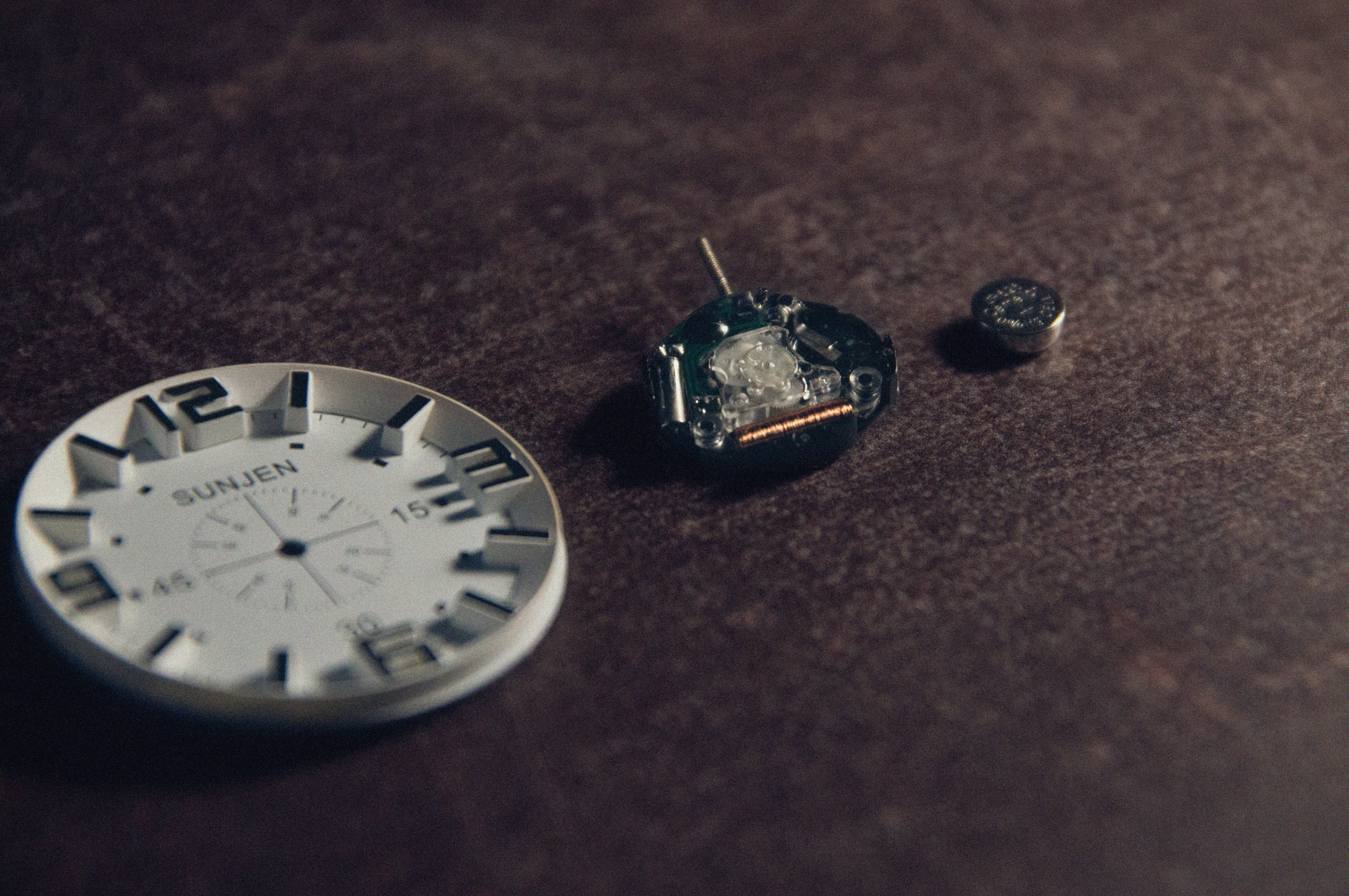 Close-up image of one white watch face, movement, and battery on a brown surface